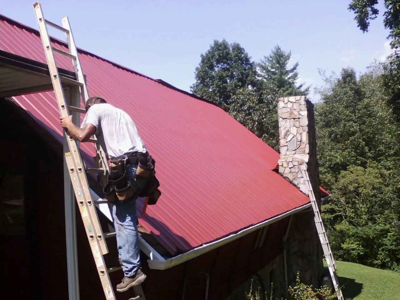 Roofer on a ladder beside a red metal roof and stone chimney — Soffit & Fascia Repair by Roofing Brothers in Wrens